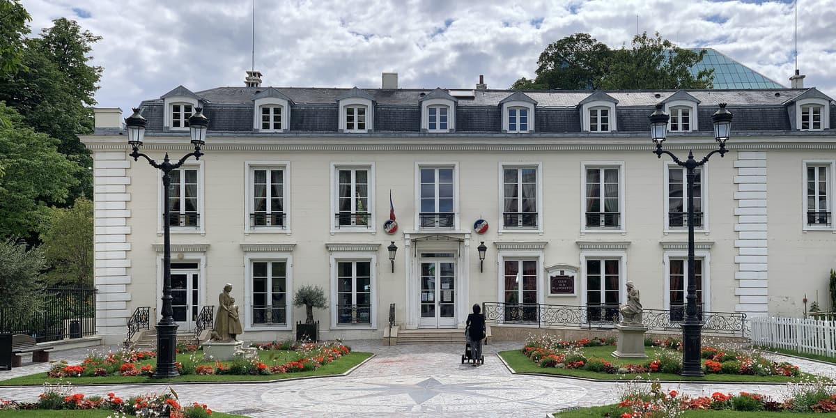 Bâtiment classique à deux niveaux et combles mansardés, façade claire symétrique entre deux chaînes d'angle, jardin à la française devant.