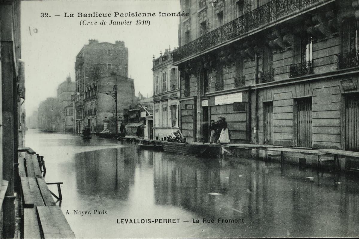Carte postale en noir et blanc : une rue d'immeubles envahie par l'eau, des passerelles de planches le long des façades.