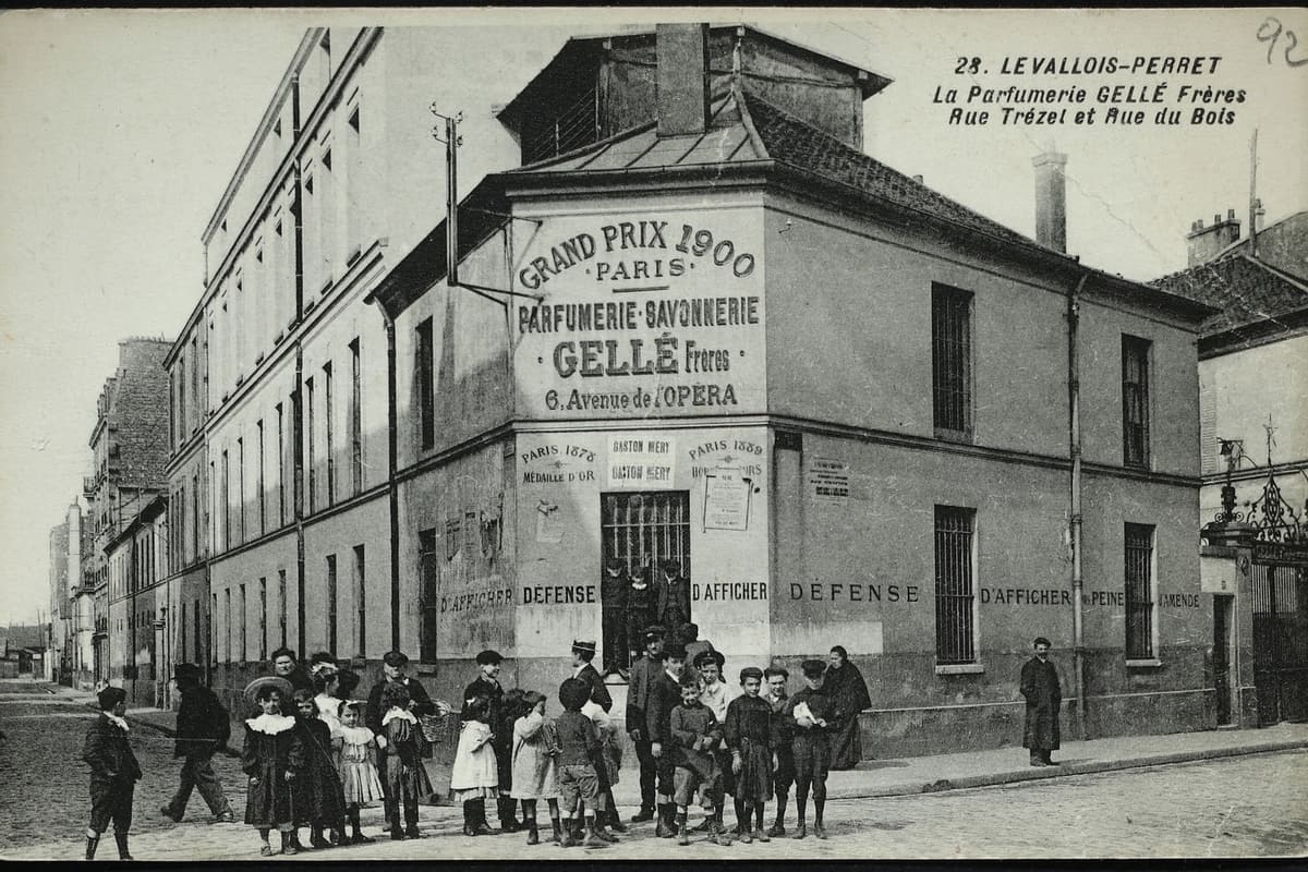 Carte postale ancienne : bâtiment d'angle de la parfumerie Gellé frères couvert d'inscriptions publicitaires, longue aile sur la rue, enfants et passants devant l'entrée.