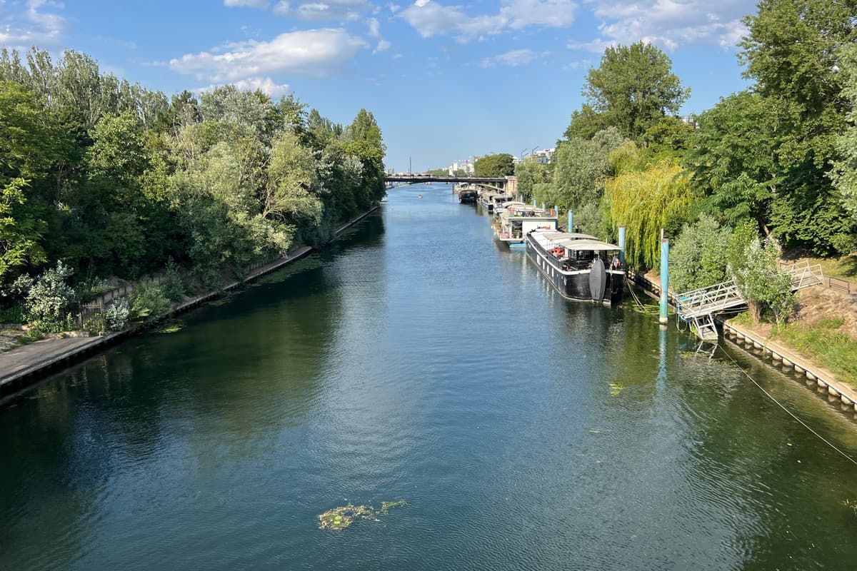 Un bras de la Seine entre deux rives boisées, des péniches amarrées à droite, un pont au loin.