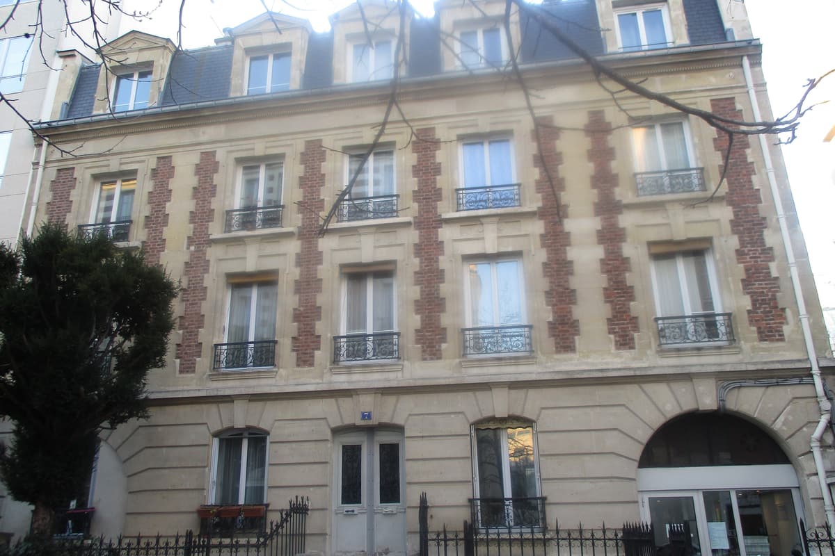 Façade d'un immeuble de 1885 : rez-de-chaussée en pierre, étages de brique et pierre, lucarnes à fronton, grille sur un muret devant une courette.